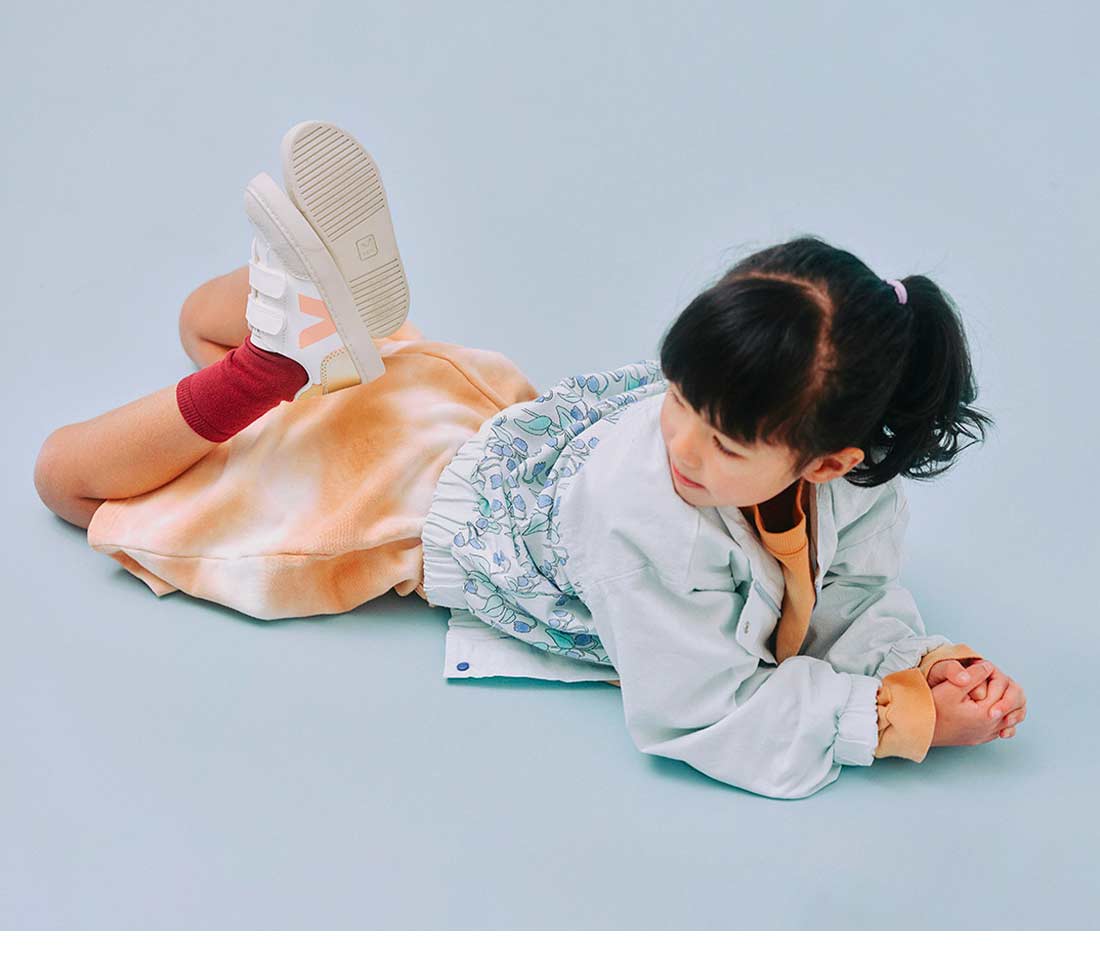 Chica tumbada en el suelo con fondo azul cielo, calzando zapatillas V-12 blancas multico platino